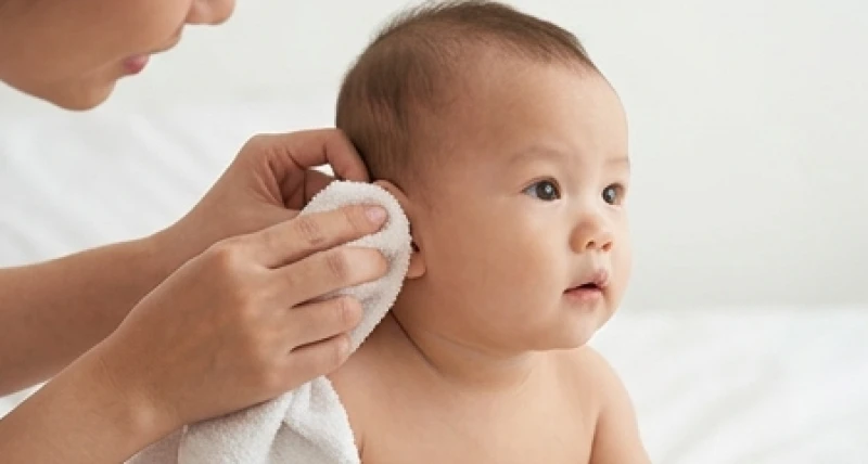 Bàn tay người lớn đang cẩn thận dùng khăn mềm lau vành tai cho một em bé trong không gian ấm áp
