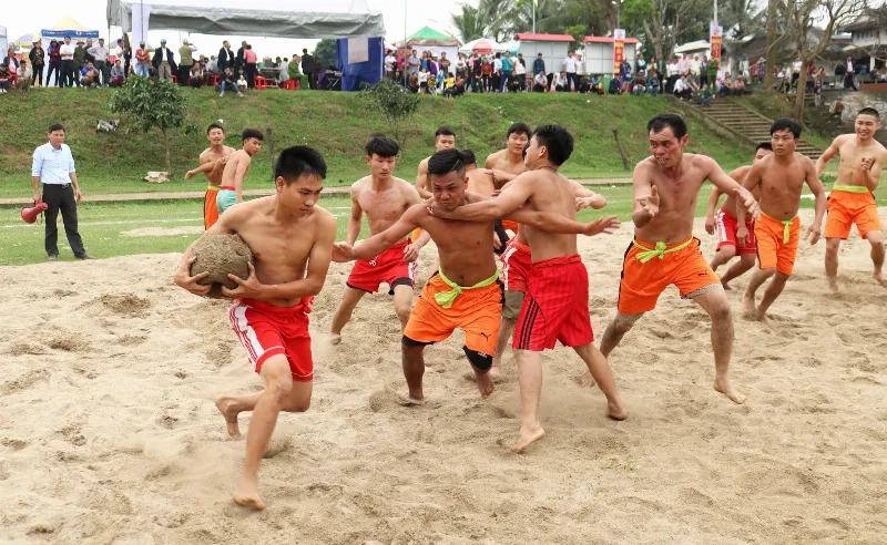 Hình ảnh minh họa lễ hội vật cù truyền thống với hai đội nam thanh niên đang tranh cướp quả cù bằng gỗ trên sân đất, thu hút đông đảo người dân đến xem và cổ vũ