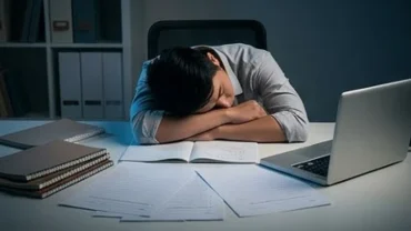 Một người trưởng thành gục đầu mệt mỏi bên bàn làm việc đầy đề thi giải thích việc mơ thấy đi học lại