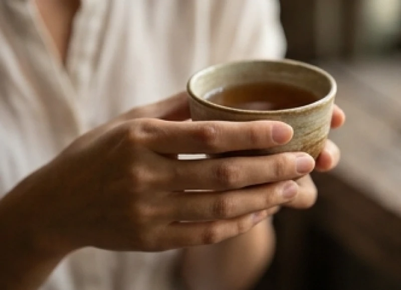 Bàn tay thon gọn với ngón cái mảnh khảnh đang nhẹ nhàng cầm một tách trà gốm sứ mộc mạc
