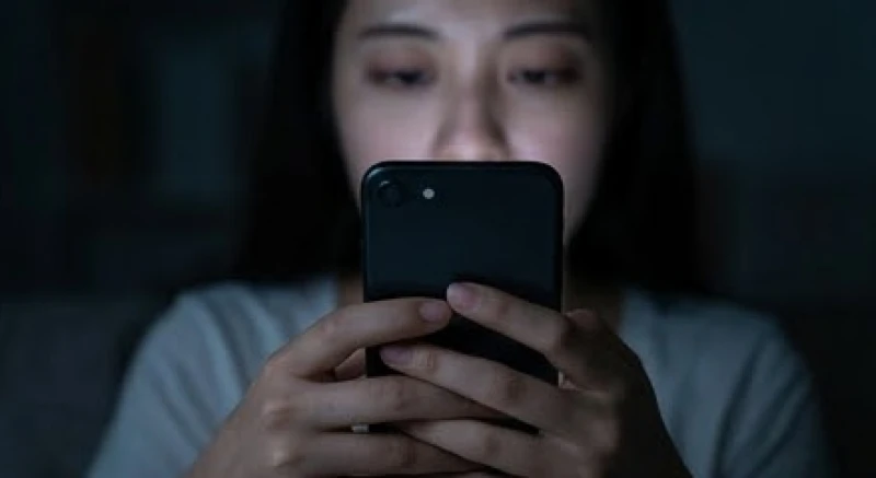 Bàn tay cầm điện thoại lướt mạng xã hội trong bóng tối với ánh sáng màn hình hắt lên gương mặt đầy mệt mỏi